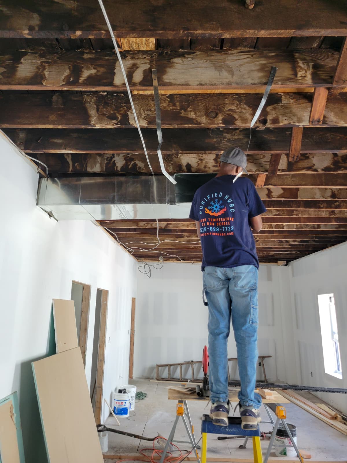Purified HVAC technician routing supply ductwork through open ceiling during gut renovation